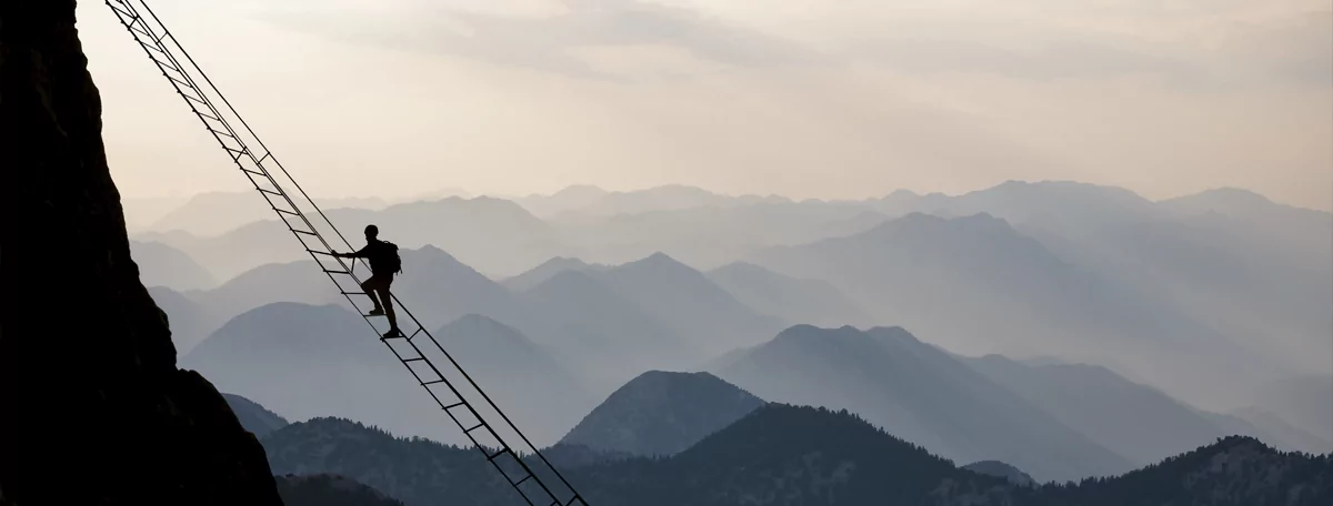 silhuette of a person hiking of a steep ladder leading to the top of a vertical cliff-face